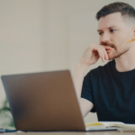 Serious bearded man student enjoys studying process has thoughtful expression works freelance dressed in casual black t shirt writes down notes uses modern laptop computer poses in coworking space