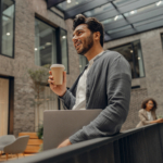 Smiling male freelancer with laptop is standing on coworking background and drinking coffee