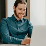 Smiling young male office worker typing on keyboard and looking at laptop screen in office.