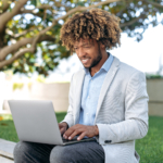 Positive attractive successful Brazilian or African American manager, freelance, stylishly elegantly dressed, male business person, sitting outdoors, working in a laptop on a project, smiling friendly